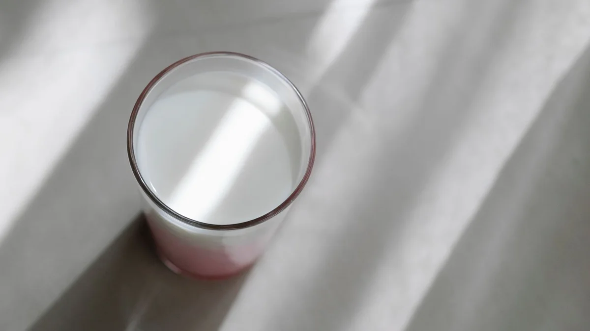A glass of milk on a pale surface, crossed by bands of soft daylight.